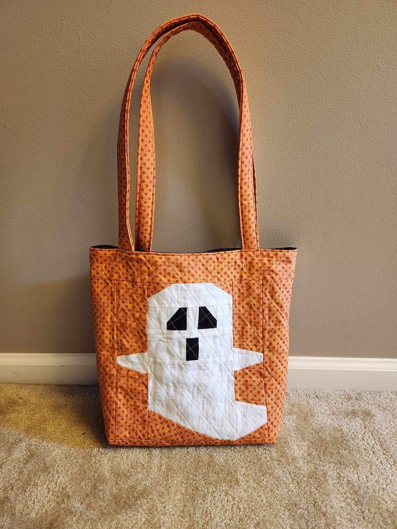 Op de afbeelding: Oranje en witte Halloween-tas met een spookontwerp. De tas heeft een wit spook met zwarte ogen en een mond op een oranje achtergrond met witte stippen.