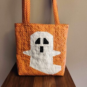 Op de afbeelding: Oranje en witte Halloween-tas met een spookontwerp. De tas heeft oranje stippen en een wit spook met zwarte ogen en een mond.