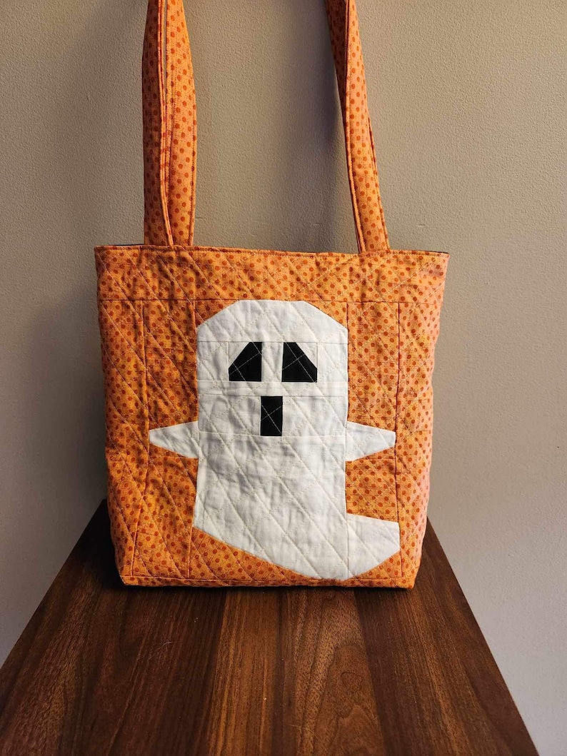 Op de afbeelding: Oranje en witte Halloween-tas met een spookontwerp. De tas heeft oranje stippen en een wit spook met zwarte ogen en een mond.