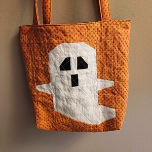 Op de afbeelding: Oranje en witte quilted tote bag met een spookontwerp. Het spook is wit met zwarte ogen en een mond.