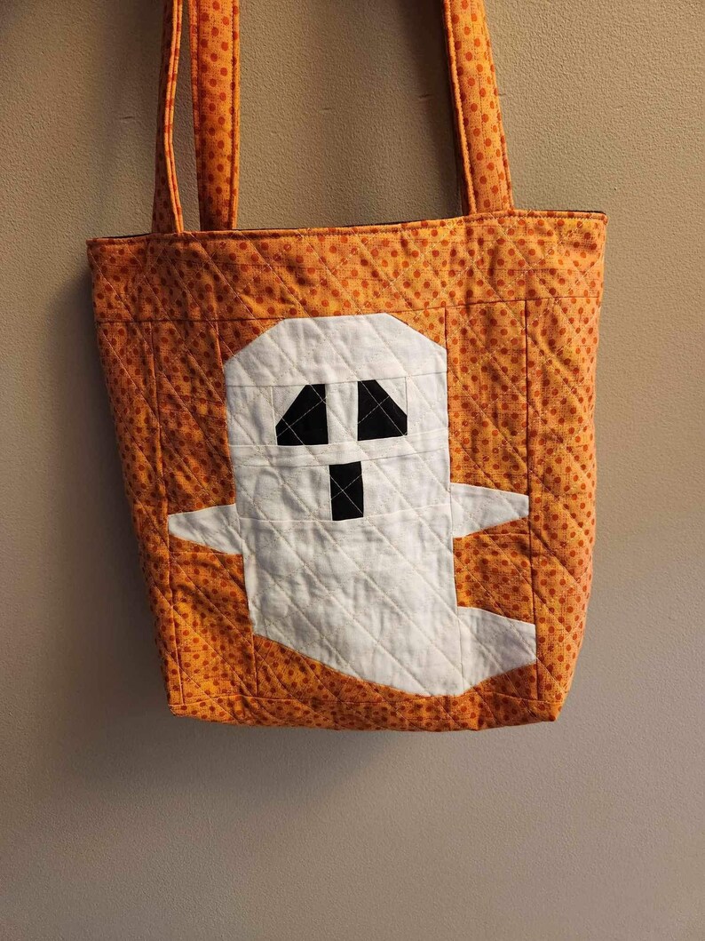 Op de afbeelding: Oranje en witte quilted tote bag met een spookontwerp. Het spook is wit met zwarte ogen en een mond.