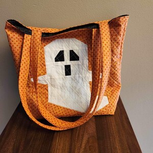 Op de afbeelding: Oranje en witte quilted tote bag met een spookontwerp. Het spook is gemaakt van witte stof met zwarte ogen en een mond.