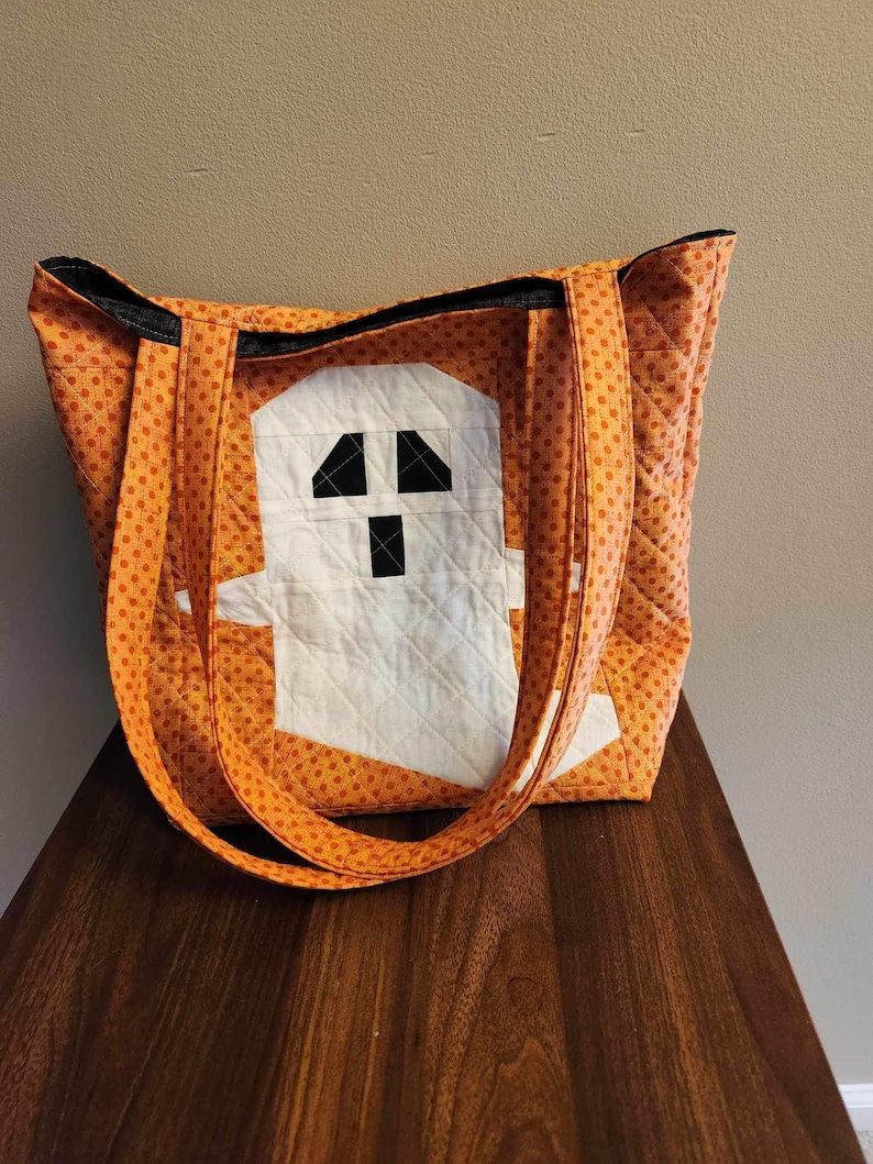 Op de afbeelding: Oranje en witte quilted tote bag met een spookontwerp. Het spook is gemaakt van witte stof met zwarte ogen en een mond.
