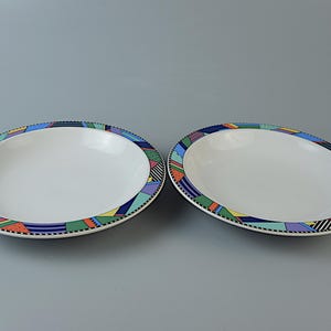 Könnte beinhalten: Zwei ovale Servierschalen aus weißer Keramik. Jede Schale hat einen farbenfrohen geometrischen Rand mit blauen, grünen, orangefarbenen und violetten Formen. Die Schalen stehen vor einem grauen Hintergrund.