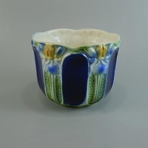 Op de afbeelding: Een keramische plantenpot met een diepblauwe glazuurlaag en verticale panelen. De buitenkant is versierd met reliëf bloemmotieven in tinten groen, geel en blauw. De rand heeft een geschulpte vorm en de binnenkant is glad en gebroken wit.