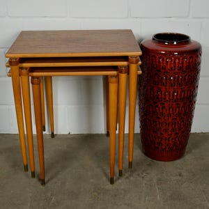 May include: A set of three light brown wooden nesting tables with tapered legs and brass-toned feet, positioned next to a tall, cylindrical, dark red ceramic floor vase featuring a textured, repeating embossed pattern across its exterior surface.