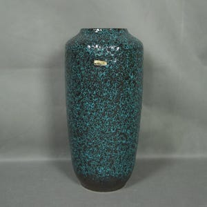 Könnte beinhalten: Hohe, zylindrische Vase mit gesprenkelter türkisblauer und schwarzer Glasur. Die Keramikvase hat einen dunkelbraunen Sockel und einen leicht ausgestellten Rand. Ein kleines Etikett ist angebracht. Geeignet für Blumenarrangements.