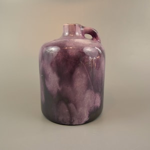 Puede incluir: Una jarra de cerámica morada con asa. La jarra tiene un esmalte jaspeado con tonos morados y negros.