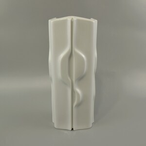 Könnte beinhalten: Weiße Keramikvase mit einem einzigartigen, abstrakten Design. Die Vase hat eine hohe, rechteckige Form mit einer Reihe von geschwungenen Linien und Vertiefungen.