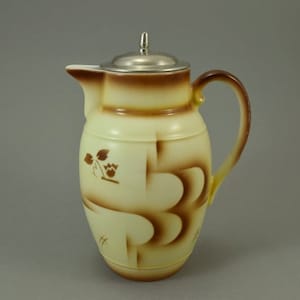 Pode incluir: Uma jarra de cerâmica de cor creme com uma tampa marrom e desenhos geométricos marrons. A jarra tem uma tampa prateada e uma alça.