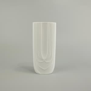 Op de afbeelding: Een hoge, witte keramische vaas met een rechthoekige vorm en een decoratief, verticaal geribbeld ontwerp. De vaas heeft een gestileerd, gebogen patroon aan de basis, wat een vleugje elegantie toevoegt aan de minimalistische esthetiek. Geschikt voor interieurdecoratie.