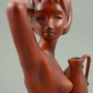 May include: A red ceramic sculpture of a woman holding a pitcher. The woman is nude and has a long, flowing hairstyle. The pitcher is brown and has a handle.