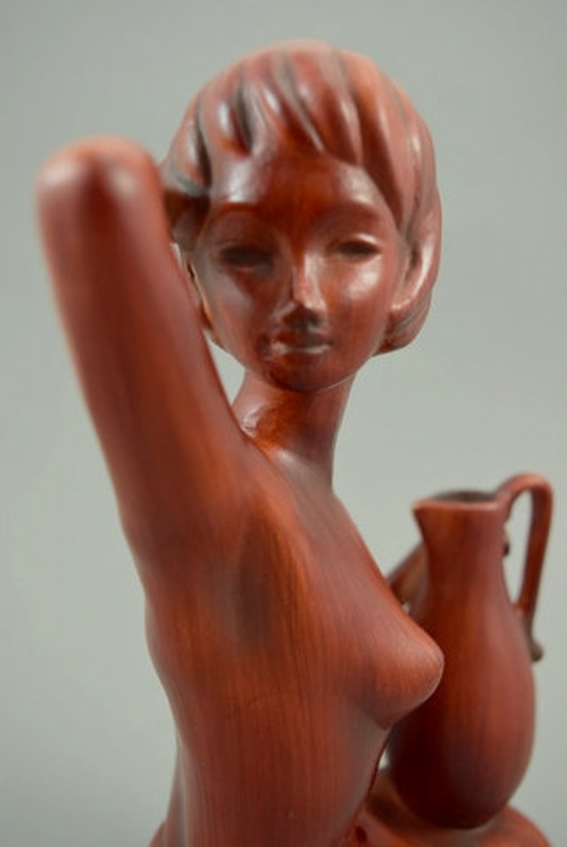 May include: A red ceramic sculpture of a woman holding a pitcher. The woman is nude and has a long, flowing hairstyle. The pitcher is brown and has a handle.