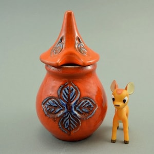 Könnte beinhalten: Eine rote Keramikdose mit einem blauen und weißen Blumendesign und einem passenden Deckel in Form eines Vogelkopfes. Im Vordergrund befindet sich eine kleine orangefarbene Plastikhirschfigur.