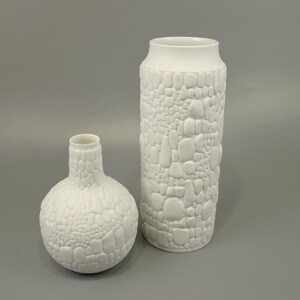Könnte beinhalten: Zwei weiße Keramikvasen mit einem strukturierten, kieselsteinartigen Design. Die Vase links ist rund und die Vase rechts ist hoch und zylindrisch.