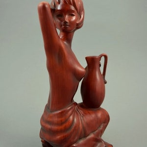 May include: A wooden sculpture of a woman holding a pitcher. The sculpture is made of a reddish-brown wood and is in a seated position. The woman is wearing a simple garment and has a serene expression on her face.