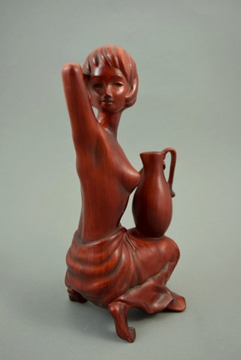 May include: A wooden sculpture of a woman holding a pitcher. The sculpture is made of a reddish-brown wood and is in a seated position. The woman is wearing a simple garment and has a serene expression on her face.