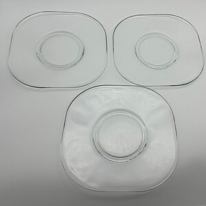 Puede incluir: Tres platos de vidrio transparente. Cada plato tiene forma cuadrada con esquinas redondeadas y una hendidura circular en el centro. Los platos están dispuestos en grupo, uno centrado y dos encima.