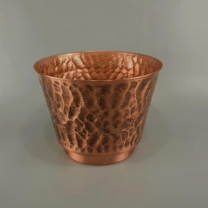Può includere: Un vaso in rame martellato con superficie strutturata. Il recipiente color rame ha un'ampia apertura e una forma leggermente conica. La texture martellata conferisce al vaso un aspetto rustico e artigianale. Ideale per uso interno o esterno.