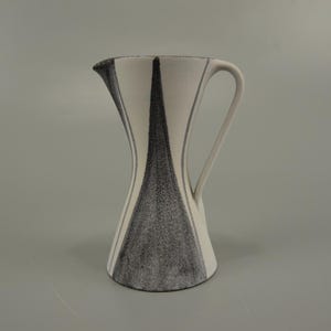 May include: A white ceramic pitcher with a black and white geometric design. The pitcher has a wide base and a narrow neck, with a handle on one side.