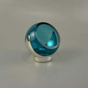Può includere: Un globo di vetro color teal con base e dettagli argentati. Il globo ha un'apertura asimmetrica insolita sul lato. L'interno è visibile attraverso l'apertura. È un oggetto decorativo.