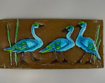 May include: A ceramic tile with three blue and green birds with long necks standing on a brown background with green reeds.