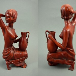 May include: A wooden sculpture of a woman holding a pitcher. The sculpture is made of a reddish-brown wood and is in a seated position. The woman is wearing a simple garment and has her hair pulled back.