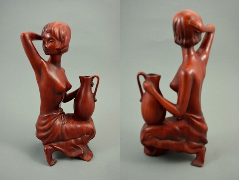 May include: A wooden sculpture of a woman holding a pitcher. The sculpture is made of a reddish-brown wood and is in a seated position. The woman is wearing a simple garment and has her hair pulled back.