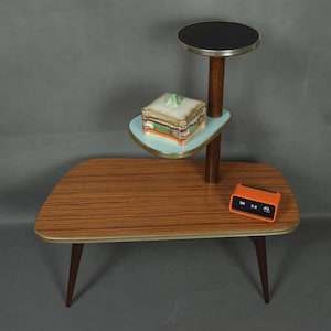Op de afbeelding: Een plantenstandaard uit het midden van de eeuw met een houten tafelblad en poten. Het heeft een lichtblauwe plank, een zwart blad en een oranje klok. De standaard is ongeveer 76 cm lang.