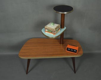 May include: A mid-century modern plant stand with a wooden tabletop and legs. It features a light blue shelf, a black top, and an orange clock. The stand is approximately 30 inches long.
