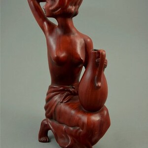 May include: A wooden sculpture of a woman holding a vase. The sculpture is made of a reddish-brown wood and is in a seated position. The woman is nude and has her hair pulled back. The vase is also made of wood and is in the shape of a gourd.
