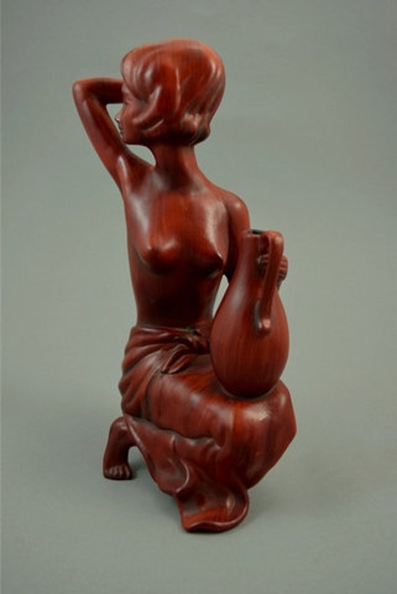 May include: A wooden sculpture of a woman holding a vase. The sculpture is made of a reddish-brown wood and is in a seated position. The woman is nude and has her hair pulled back. The vase is also made of wood and is in the shape of a gourd.