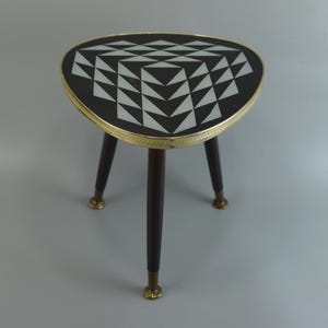 May include: A three-legged side table with a triangular top featuring a black and white geometric triangle pattern. The tabletop is edged with a gold-toned metal band. The three tapered legs are dark brown wood with gold-toned metal feet.