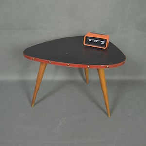 Op de afbeelding: Een driehoekige bijzettafel in mid-century stijl met een zwart blad, rode rand en houten poten. Een oranje retro wekker staat op de tafel. De tafel heeft drie poten.