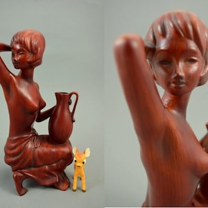May include: A wooden sculpture of a woman holding a pitcher. The sculpture is painted in a reddish-brown color and has a detailed design. The woman is wearing a simple garment and has a deer figurine at her feet.