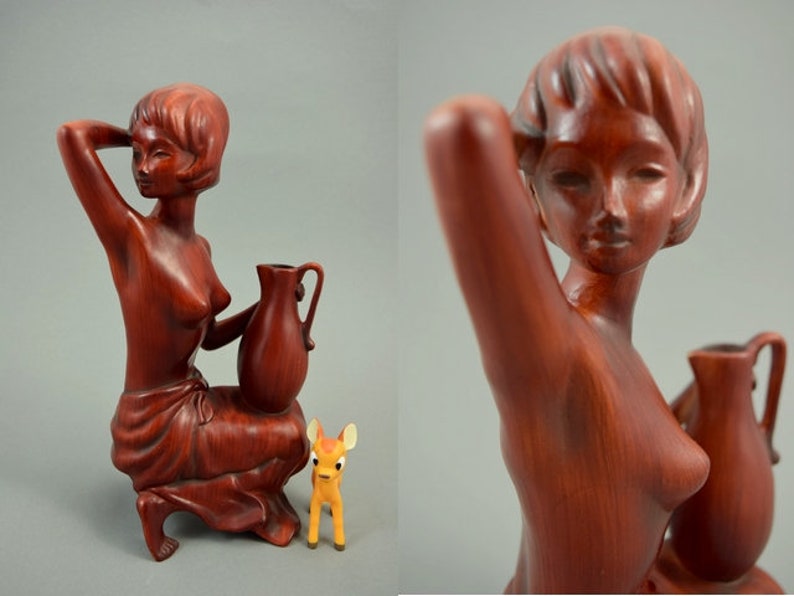 May include: A wooden sculpture of a woman holding a pitcher. The sculpture is painted in a reddish-brown color and has a detailed design. The woman is wearing a simple garment and has a deer figurine at her feet.