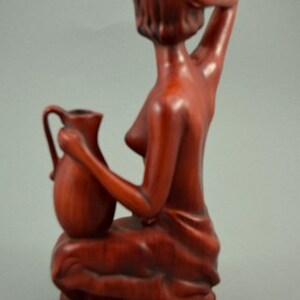 May include: A wooden sculpture of a woman holding a pitcher. The sculpture is made of a reddish-brown wood and is in a seated position. The woman is wearing a simple garment and has her hair pulled back.