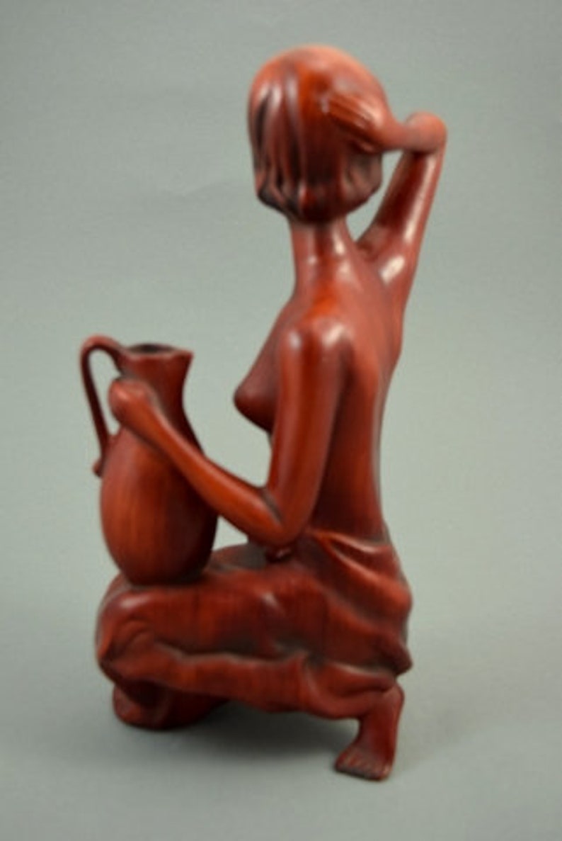 May include: A wooden sculpture of a woman holding a pitcher. The sculpture is made of a reddish-brown wood and is in a seated position. The woman is wearing a simple garment and has her hair pulled back.