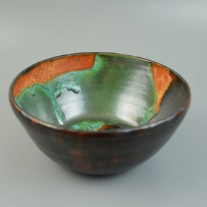 Op de afbeelding: Een handgemaakte keramische kom met een donkerbruine buitenkant en een levendig interieur. De binnenkant heeft een mix van groene en turquoise tinten, geaccentueerd door een oranje band bij de rand. Deze decoratieve kom is ideaal om te serveren of tentoon te stellen.