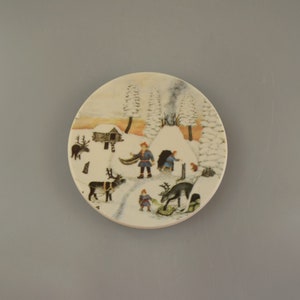 May include: A white ceramic plate with a winter scene featuring a small cabin, trees, and people in a snowy landscape. The plate has a brown rim and a white background.