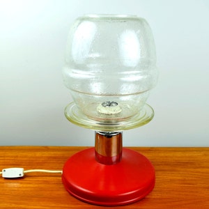Może przedstawiać: Czerwona lampa stołowa z chromowanym podstawą i przezroczystym kloszem. Klosz ma fakturowaną powierzchnię i okrągłą podstawę.