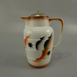 Pode incluir: Uma jarra de cerâmica branca com tampa prateada e um design geométrico em preto, laranja e branco.