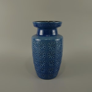 Puede incluir: Un jarrón de cerámica azul con un patrón geométrico. El jarrón tiene un cuerpo redondeado y una abertura ligeramente acampanada.