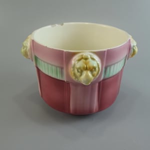 Op de afbeelding: Een keramische plantenpot met een dieprode basis en een lichter roze bovengedeelte met verticale groene strepen. De buitenkant is versierd met drie gegoten leeuwenkopmotieven in een lichtgele glazuur, de binnenkant is glad en gebroken wit.