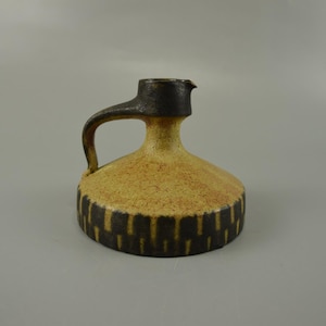 Puede incluir: Una jarra de cerámica con asa y boquilla. La jarra tiene un cuerpo redondeado con un acabado moteado amarillo y marrón. La base está decorada con rayas verticales de color marrón oscuro. El asa y la boquilla son de color marrón oscuro.