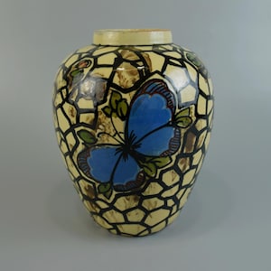 May include: A decorative ceramic vase with a cream-colored background and a black geometric pattern. A large blue butterfly with green accents is painted on the front. The vase has a rounded shape.
