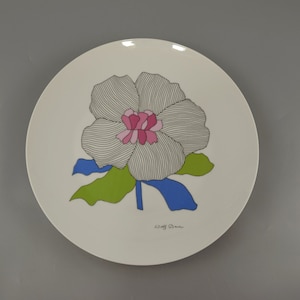 Peut inclure: Assiette en céramique blanche avec un motif de fleur stylisé en noir et blanc avec des accents roses et bleus. L'assiette a une tige et des feuilles vertes. La signature de l'artiste "Staff Dane" est imprimée au bas de l'assiette.