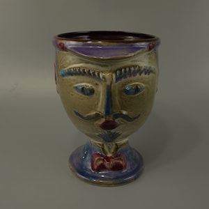 Könnte beinhalten: Ein Keramik-Kelch in Form eines Gesichts mit einer violetten, blauen und braunen Glasur. Das Gesicht hat einen Schnurrbart, Augenbrauen und eine Fliege.
