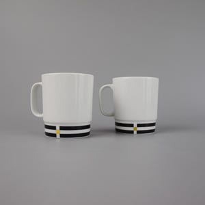 Könnte beinhalten: Zwei weiße Keramikbecher mit einem schwarz-gelb gestreiften Design am Boden. Die Becher haben ein schlichtes, modernes Design mit einem Henkel an der Seite. Der Hintergrund ist einfarbig grau.