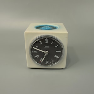 May include: A white ceramic clock with a black face and Roman numeral hour markers. The clock is in a cube shape with a blue and white design on the top.